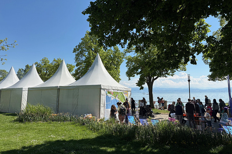 Weiße Zelte der Explore Science-Veranstaltung an der Uferpromenade in Friedrichshafen, umgeben von grünen Bäumen und Blumenbeeten. Im Vordergrund Besucher:innen an Mitmachstationen, im Hintergrund der Bodensee bei strahlendem Sonnenschein.