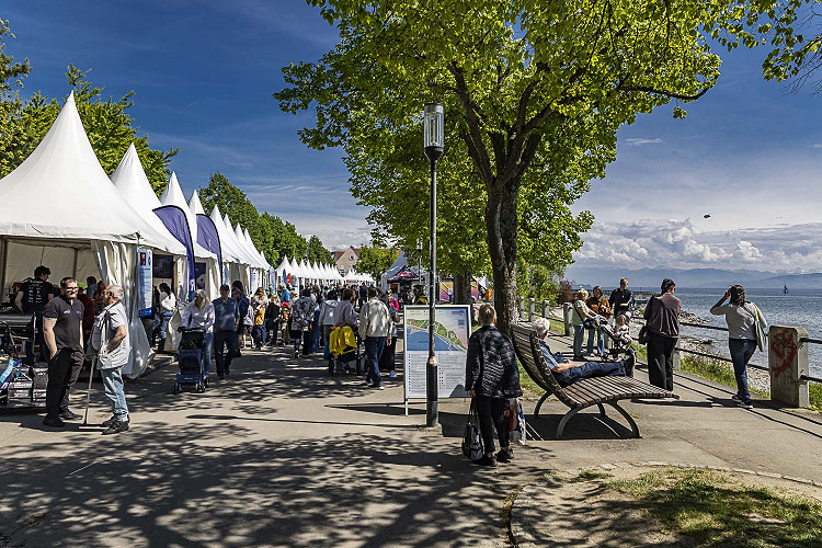 Besucherinnen und Besucher schlendern an weißen Veranstaltungszelten entlang der Uferpromenade am Bodensee.