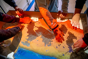 Die Jugendlichen modellieren Landschaften aus Sand, während ein Beamer sie in Echtzeit mit farbigen Höhenkarten und Wasserprojektionen überlagert – ein interaktives Experiment zum Thema Geografie und Physik.